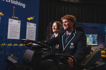 Dos estudiantes uniformados interactúan con una exhibición de simulación, sonriendo mientras operan el equipo.